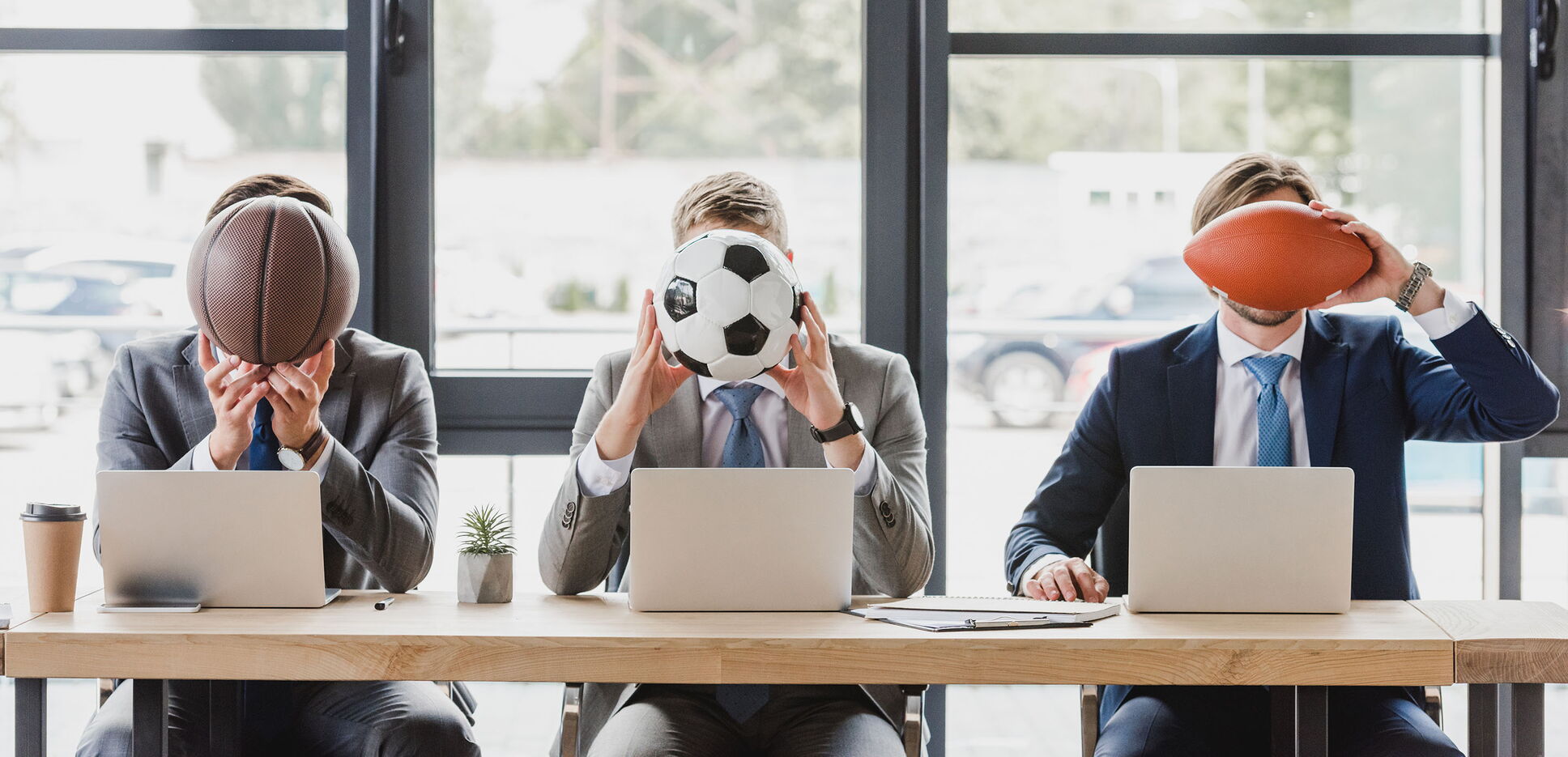 Drei Männer in Anzügen sitzen hinter ihren Laptops und halten sich Bälle aus verschiedenen Sportarten vor das Gesicht