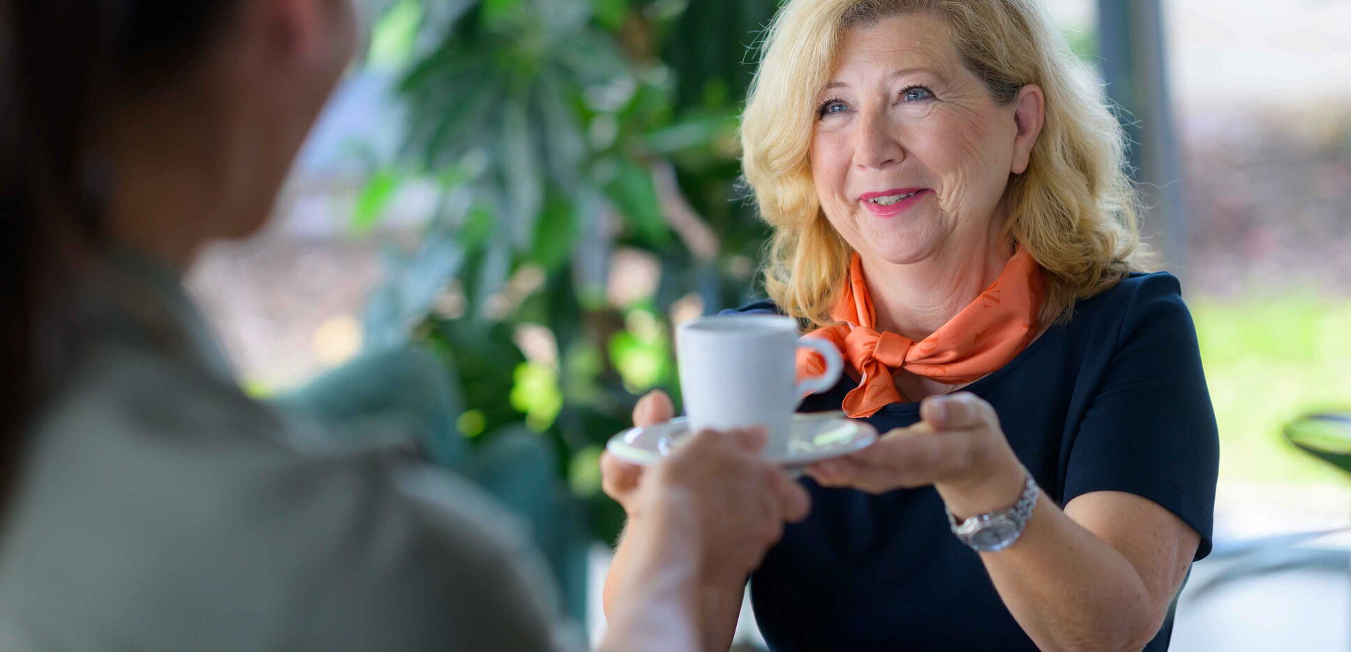Empfangsmitarbeiterin reicht einer Besucherin eine Tasse Kaffee.