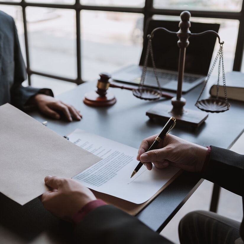 Zwei Personen sitzen sich an einem Tisch gegenüber, während eine davon ein Dokument unterschreibt, symbolisiert Korruption und Bestechung im Geschäftsumfeld, relevant für Whistleblowing-Themen.