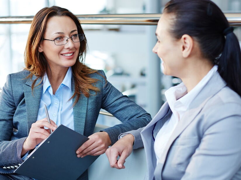 Zwei Frauen in Business-Kleidung im lockeren Vier-Augen-Gespräch, eine hält ein Klemmbrett und einen Stift – sinnbildlich für den kollegialen Austausch in der Führungskräfteentwicklung.