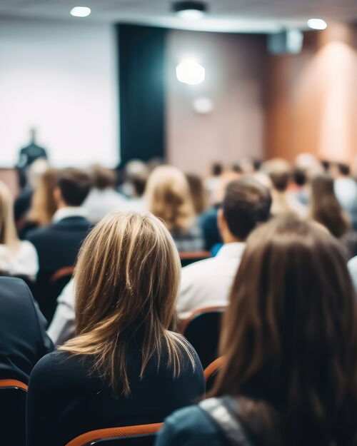 Publikum von hinten in einem Konferenzsaal blickt auf eine Bühne mit Leinwand.