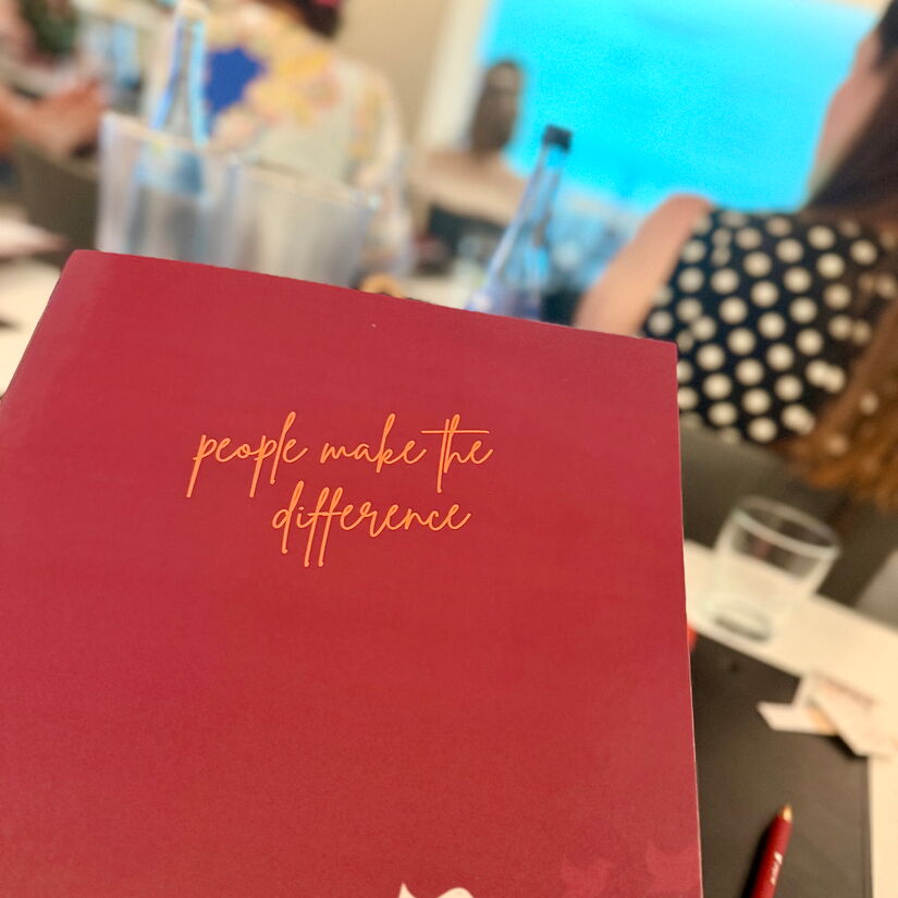 Rote Konferenzmappe mit der Aufschrift: people make the difference. Dazu peepz-Vogelsymbol, im Hintergrund Teilnehmende der Veranstaltung.