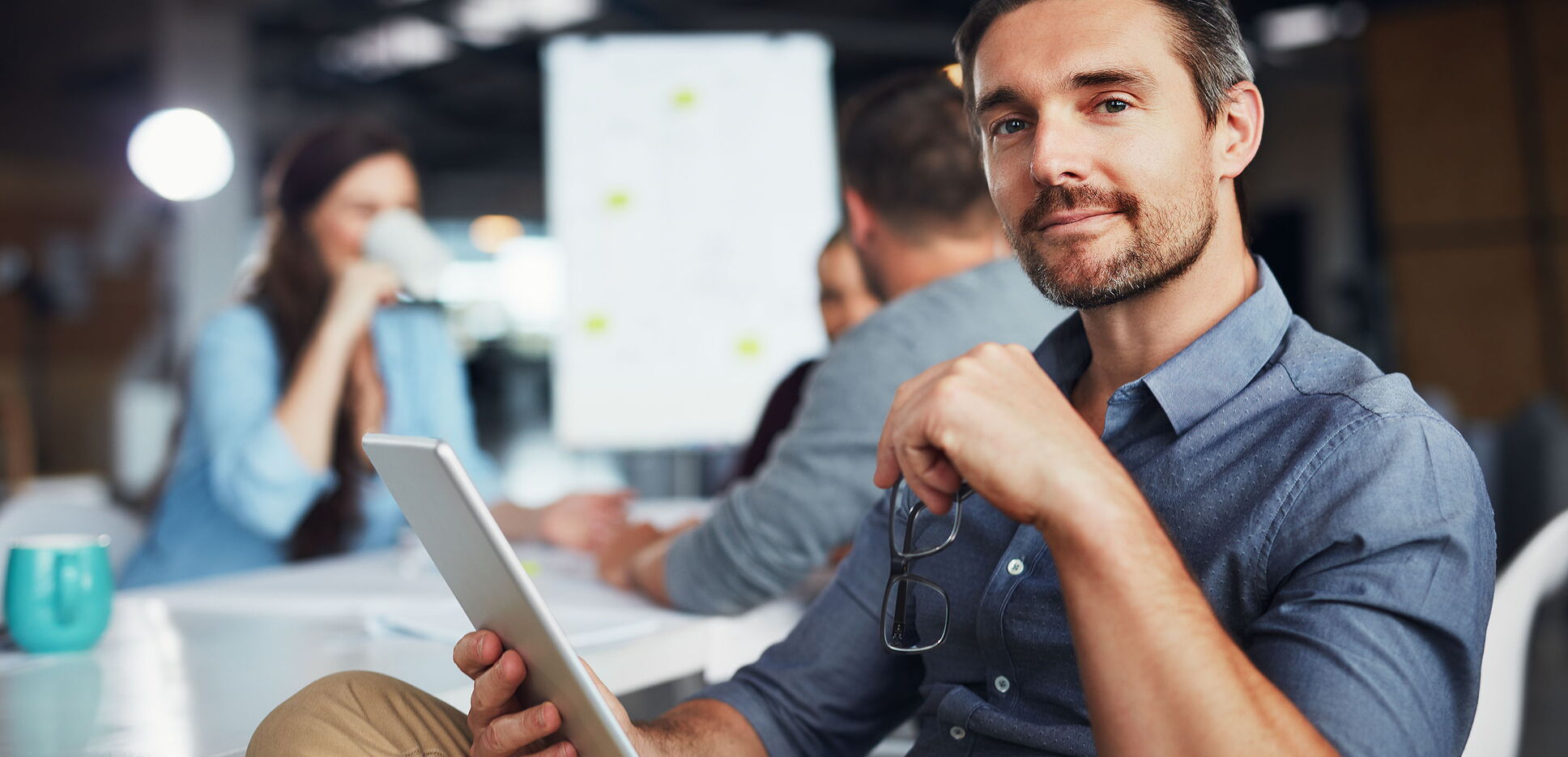 Porträt eines Mannes, der ein digitales Tablet benutzt, während im Hintergrund Kollegen arbeiten